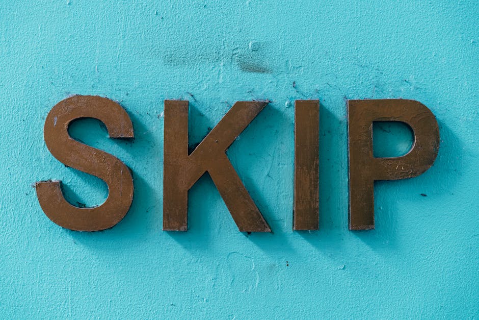 Close-up of three metallic letters spelling 'SKIP' mounted on a textured, light blue-painted wall. The letters are dark brown with a slightly weathered finish, and they are positioned in the center of