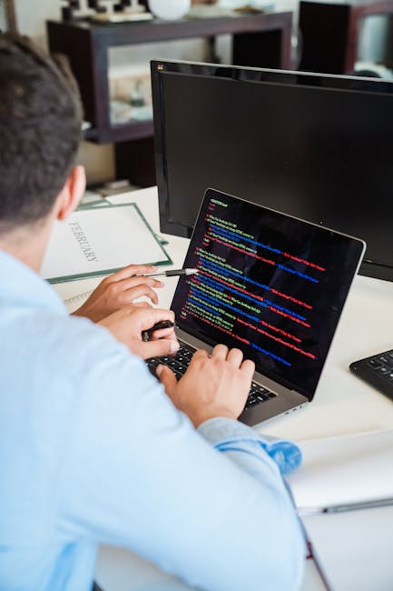 A person sitting at a desk working on a laptop displaying lines of colorful programming code on the screen. The individual is dressed in a light blue shirt and is using a wired mouse with their right